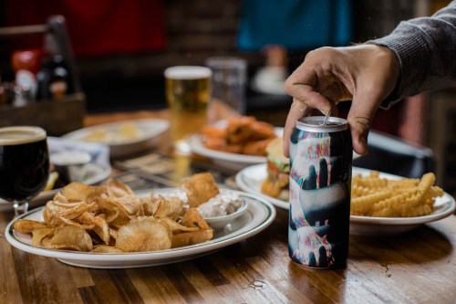 A hand opens a colorful soda can beside plates of chips, fries, and other food in a casual dining setting.