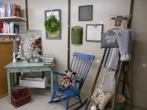 A cozy corner featuring a blue rocking chair, decorative pillows, a small tree, and a vintage-style mirror on a pegboard wall.