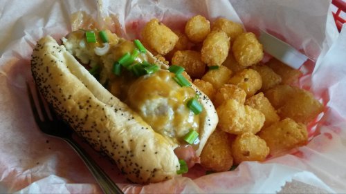 A poppy seed hot dog topped with cheese and green onions, served with a side of crispy tater tots in a red basket.