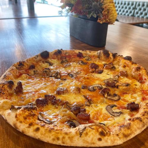 A freshly baked pizza topped with cheese, mushrooms, and crispy bacon, placed on a wooden table.