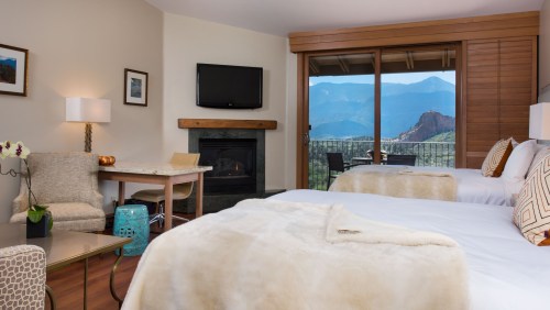 Cozy hotel room with two beds, a fireplace, and a view of mountains through large windows. Modern decor and furnishings.