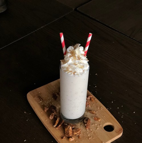 A tall glass of milkshake topped with whipped cream and crushed nuts, served with two red and white striped straws.