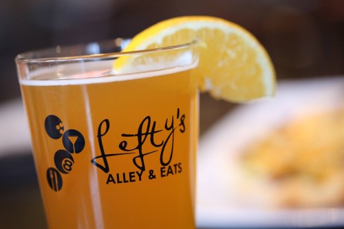 A glass of beer with a lemon slice, featuring the logo of Lefty's Alley & Eats in the foreground.