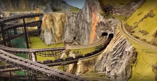 A detailed model train landscape featuring winding tracks, tunnels, and rocky terrain with lush greenery.