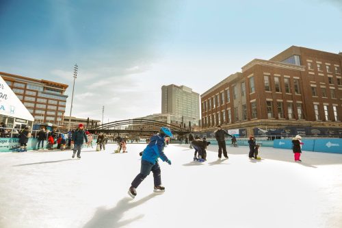 A bustling ice skating rink with people of all ages enjoying skating on a sunny day in an urban setting.
