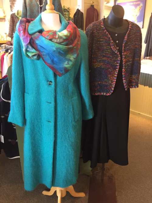 A vibrant turquoise coat with a colorful scarf, alongside a black dress and a multicolored cardigan on display.