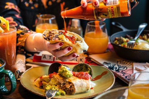 A hand drizzles hot sauce over a burrito on a plate, surrounded by drinks and other dishes on a table.