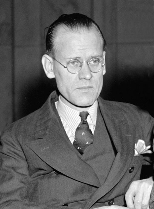 A serious man in a suit and glasses, seated with a focused expression, against a dark background.