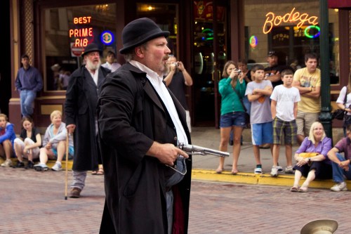 A man in period costume holds a revolver, surrounded by a crowd watching a street performance.