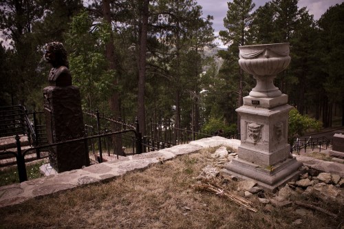A stone urn and a bust statue are set against a backdrop of trees and a distant view, on a hillside cemetery.