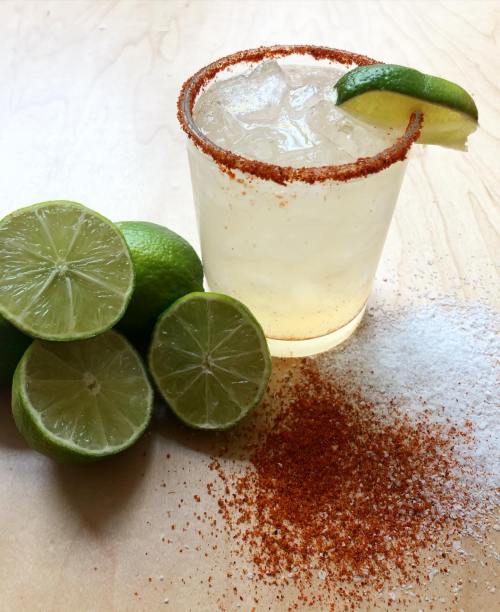 A refreshing cocktail with a lime wedge, rimmed with chili powder, beside halved limes and a sprinkle of seasoning.