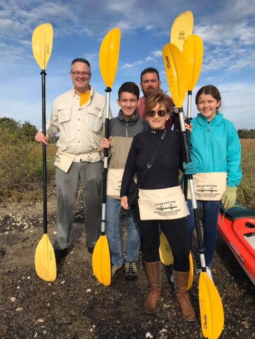 A group of five people holding yellow kayak paddles, standing on a beach with greenery in the background.