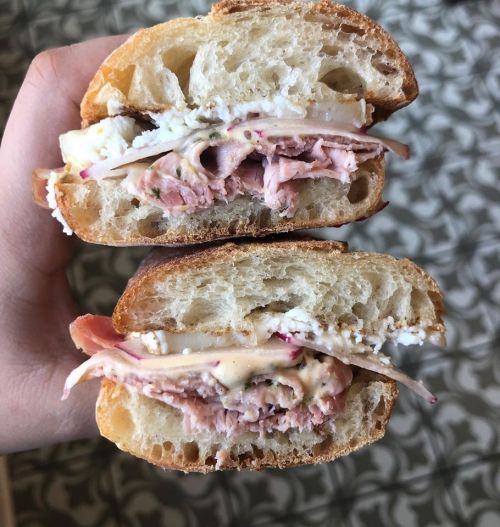 A hand holding a sandwich cut in half, revealing layers of meat, cheese, and spread between two pieces of bread.