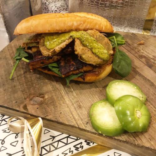 A hearty sandwich with layers of greens, fried pickles, and a green sauce, served on a wooden board with sliced cucumbers.