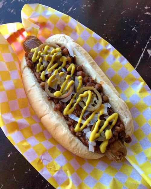 A hot dog in a bun topped with chili, onions, jalapeños, and mustard, served on a yellow checkered paper.