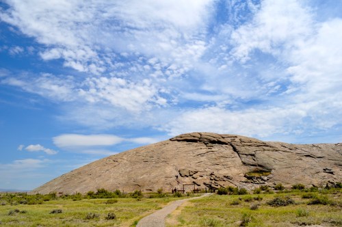 A large, smooth rock formation rises against a blue sky with scattered clouds, surrounded by green grass and a dirt path.