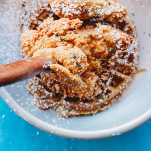 A wooden spoon scoops up crispy fried chicken pieces from a bowl, dusted with powdered sugar.