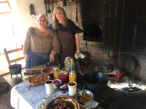 Two women in historical attire stand in a rustic kitchen, surrounded by traditional foods and cooking equipment.