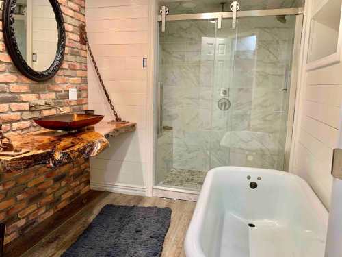 A modern bathroom featuring a brick wall, wooden sink countertop, glass shower, and a freestanding bathtub.