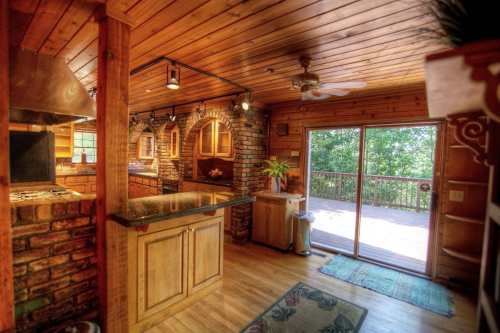 Cozy wooden kitchen with brick accents, large windows, and a sliding door leading to a deck surrounded by greenery.