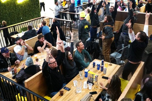 A lively crowd in a restaurant, cheering and raising their hands in excitement, with drinks on the tables.