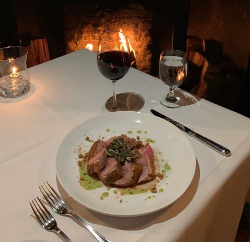A beautifully plated dish of meat with sauce, accompanied by a glass of red wine and a candlelit setting by a fireplace.