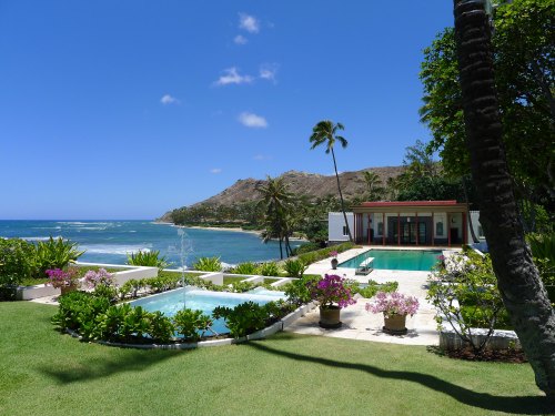 A tropical beachfront villa with lush gardens, pools, and palm trees, overlooking the ocean and mountains in the background.