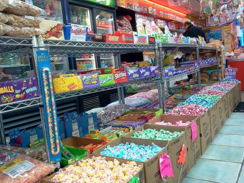 A colorful candy store aisle filled with various candies in boxes and on shelves, with a customer in the background.