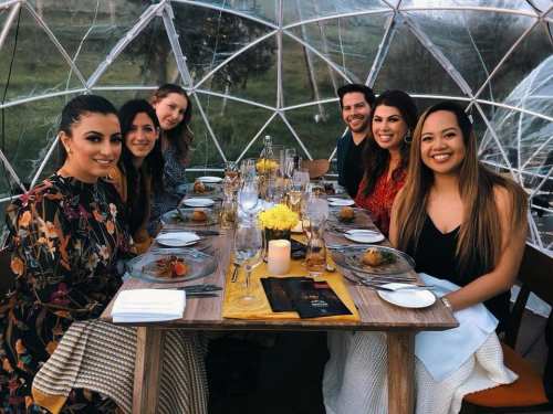A group of six people enjoying a meal together inside a transparent dome, surrounded by nature.