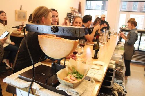 A cheese wheel melting on a stand, with patrons enjoying food and drinks at a bar in a cozy setting.