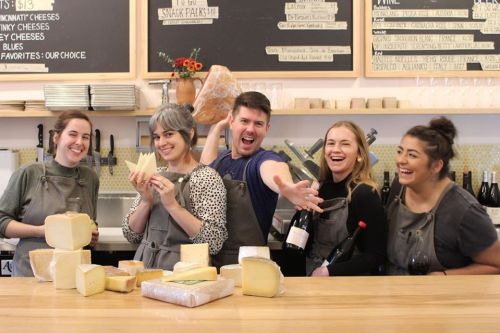 A group of five people in aprons pose joyfully with various cheeses and wine in a cozy shop setting.