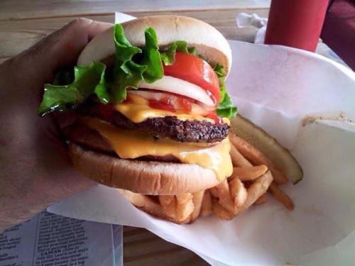 A hand holding a large cheeseburger with lettuce, tomato, and onion, accompanied by fries and a pickle.