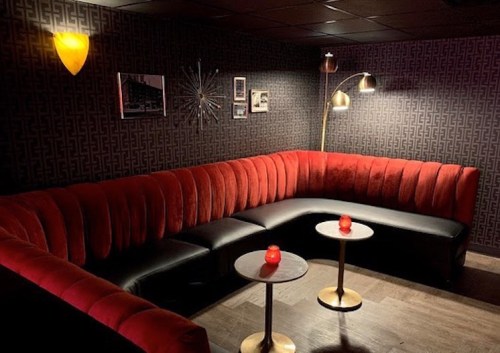 Cozy lounge with red velvet seating, black tables, and warm lighting against a patterned wall.
