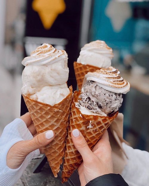 Three ice cream cones with swirled toppings held by two hands, featuring flavors like cookies and cream and vanilla.