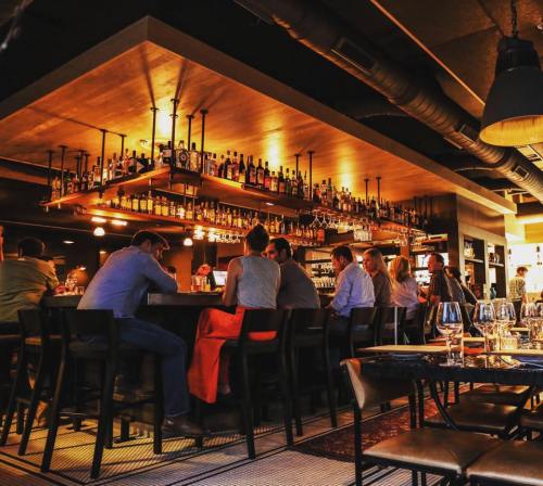 A cozy restaurant bar with patrons seated, shelves lined with bottles, and warm lighting creating an inviting atmosphere.