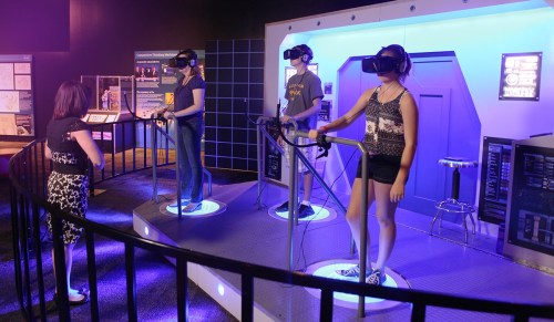 Three individuals wearing VR headsets stand on circular platforms, engaging with a virtual reality exhibit in a museum.
