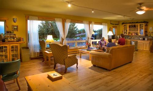 Cozy living room with wooden floors, large windows, and people gathered around a coffee table. Warm, inviting atmosphere.