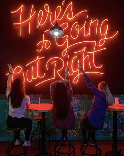 Three people sitting on bar stools, raising their hands in front of a neon sign that says, "Here's to Going Out Right."