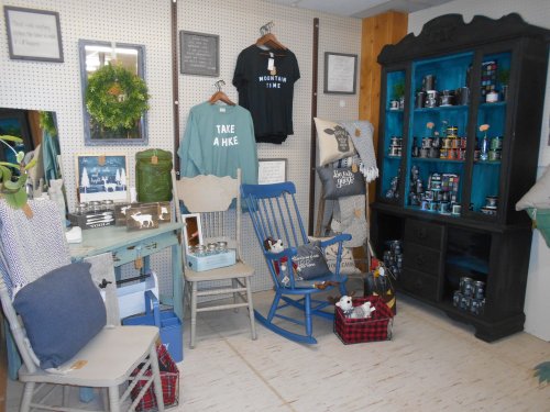 A cozy shop corner featuring rustic decor, a blue rocking chair, and shelves filled with candles and home goods.