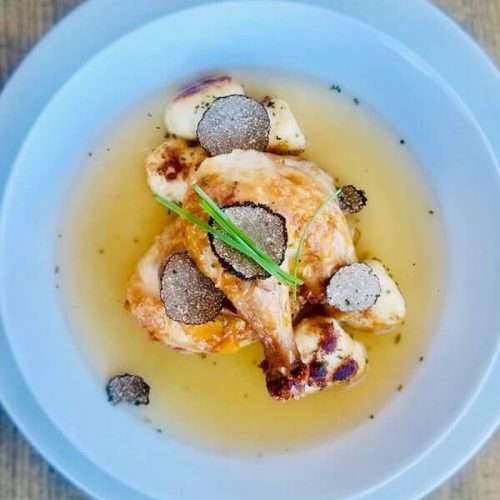 A plate of chicken with truffle slices and gnocchi in a light broth, garnished with herbs.