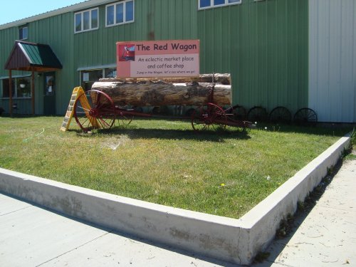 A large wooden wagon with a sign reading "The Red Wagon" sits on a grassy area outside a green building.