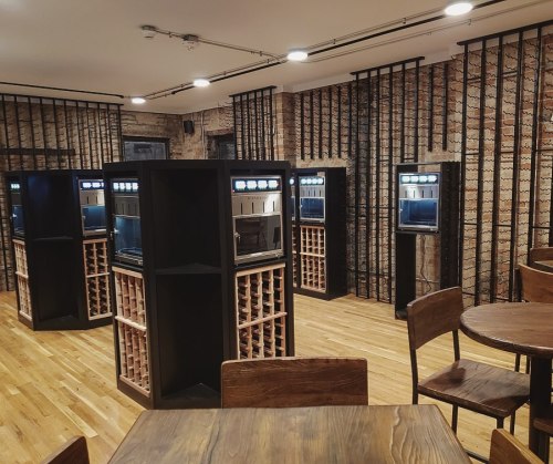 Interior of a modern space featuring wine dispensers and wooden tables, with a rustic brick wall backdrop.