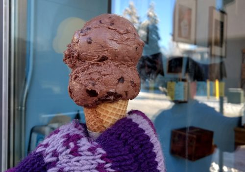 A hand holding a double scoop of chocolate ice cream in a cone, with a shop window in the background.