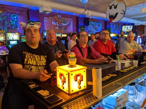 A group of gamers focused on playing video games at a retro arcade, with colorful decor and a question mark block centerpiece.