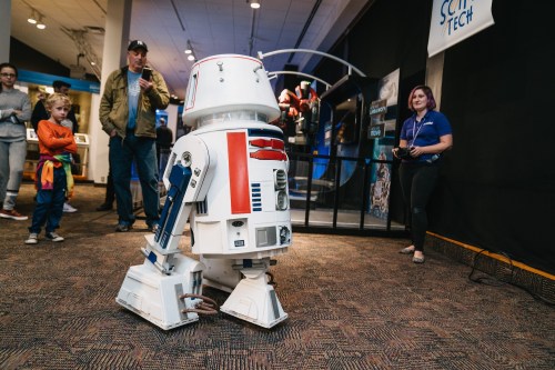 A robot resembling R2-D2 moves in a tech exhibit, with people observing and a woman controlling it nearby.