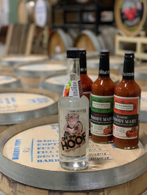 A bottle of "Off the Hoof" spirit with three bottles of Bloody Mary mix on wooden barrels in a distillery setting.