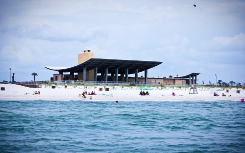 A beach scene featuring a modern building on white sand, with people relaxing and swimming in the turquoise water.