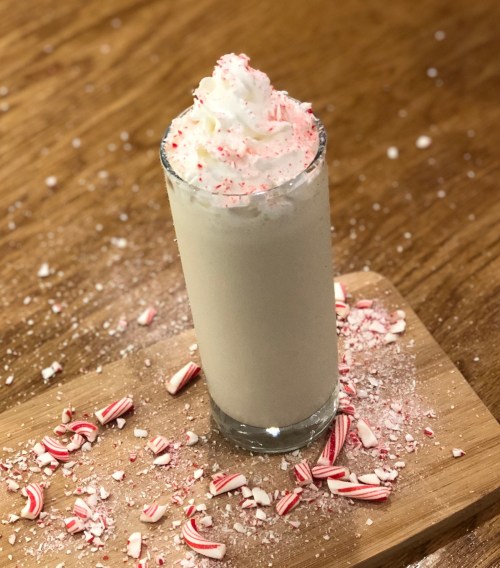 A tall glass of creamy white drink topped with whipped cream and crushed peppermint, surrounded by candy cane pieces.