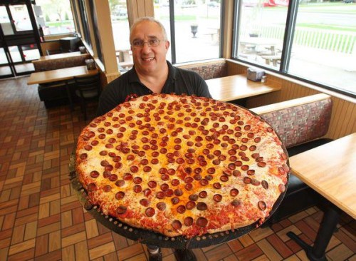 A man proudly holds a large pepperoni pizza in a restaurant with empty tables in the background.