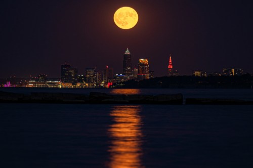 A full moon rises over a city skyline at night, reflecting on the water with colorful lights from buildings.